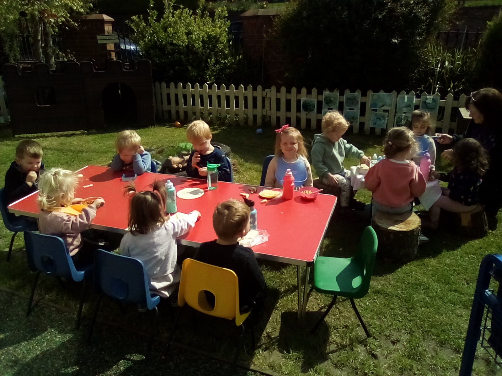 Children seated around a red table in a sunny outdoor setting, eating and interacting.