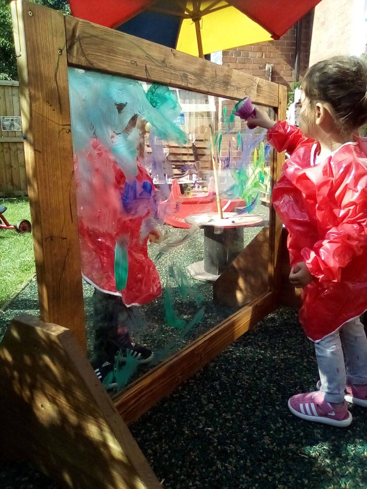 Child in red smock painting on a clear surface, outdoors. Frame made of wood; colorful paint visible.
