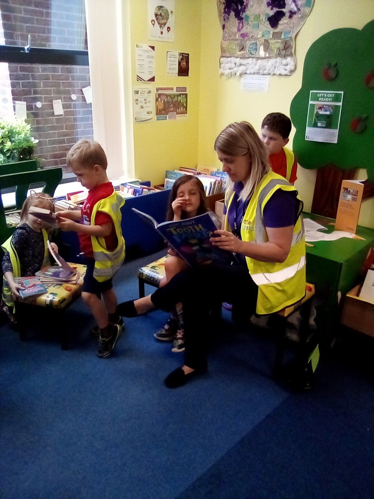 Children and adult reading a book in a classroom; they wear safety vests. Blue carpet, yellow walls, and a tree decoration.