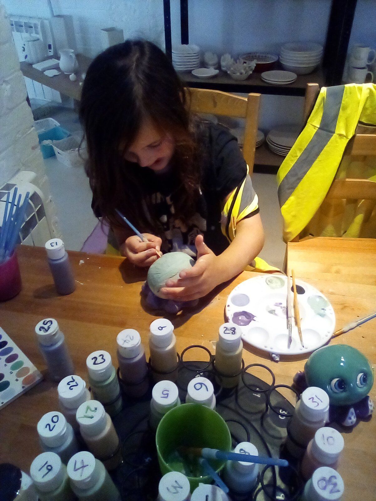 Young person paints a ceramic item at a table with paint bottles and a palette in an art studio.