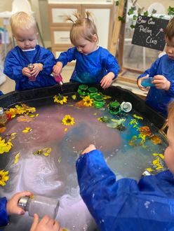 Children in blue smocks playing with colorful water and flowers in a tray.
