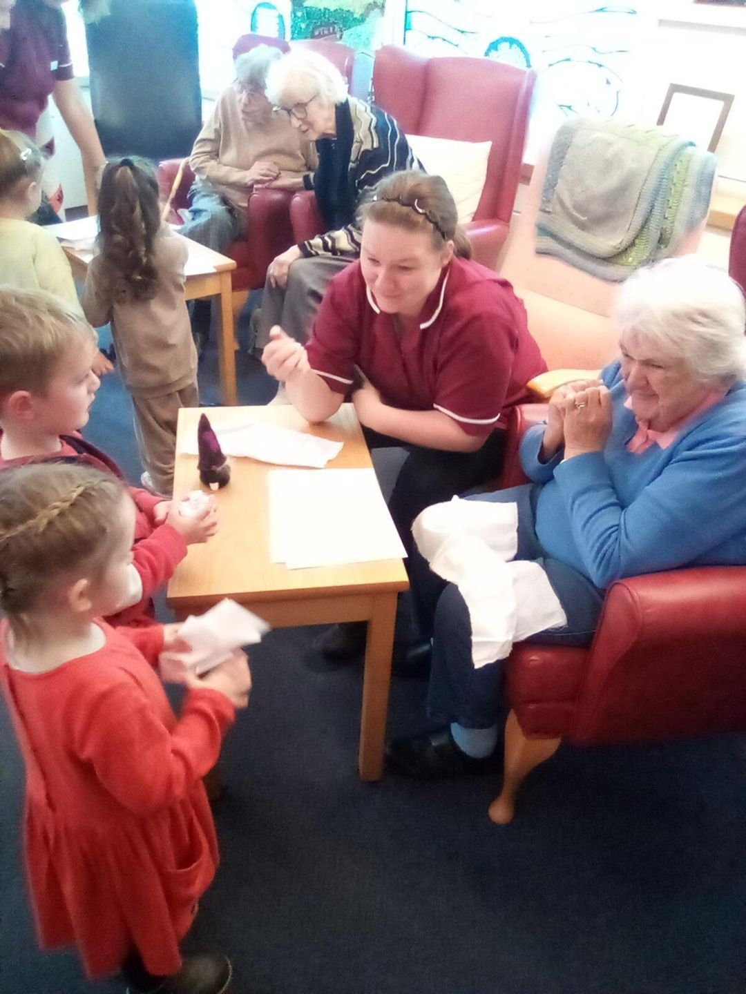 Children and a caregiver interact with an elderly woman at a table. Others relax in the background.