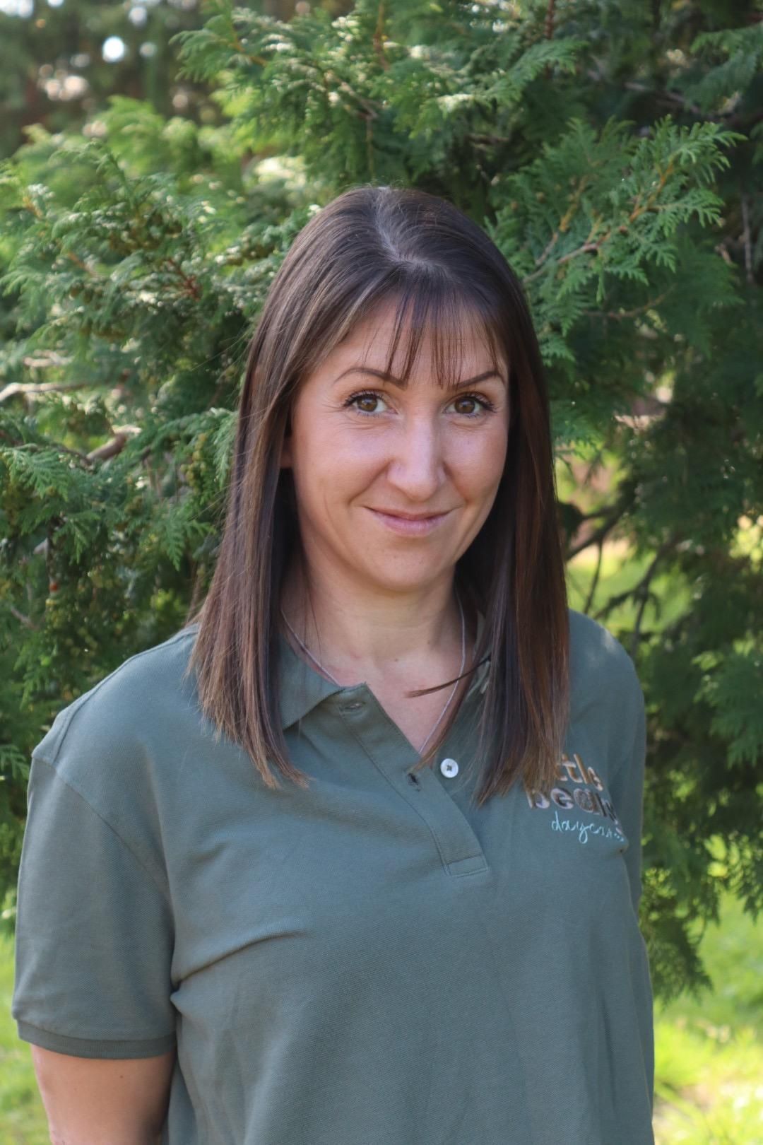 Woman in green polo shirt, smiling, standing outdoors in front of green foliage.