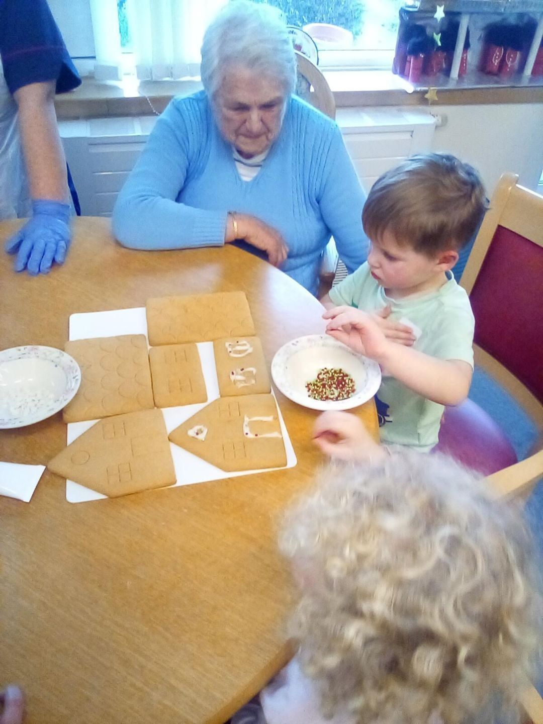 A woman and child decorate a gingerbread house at a table; another person has gloved hands.