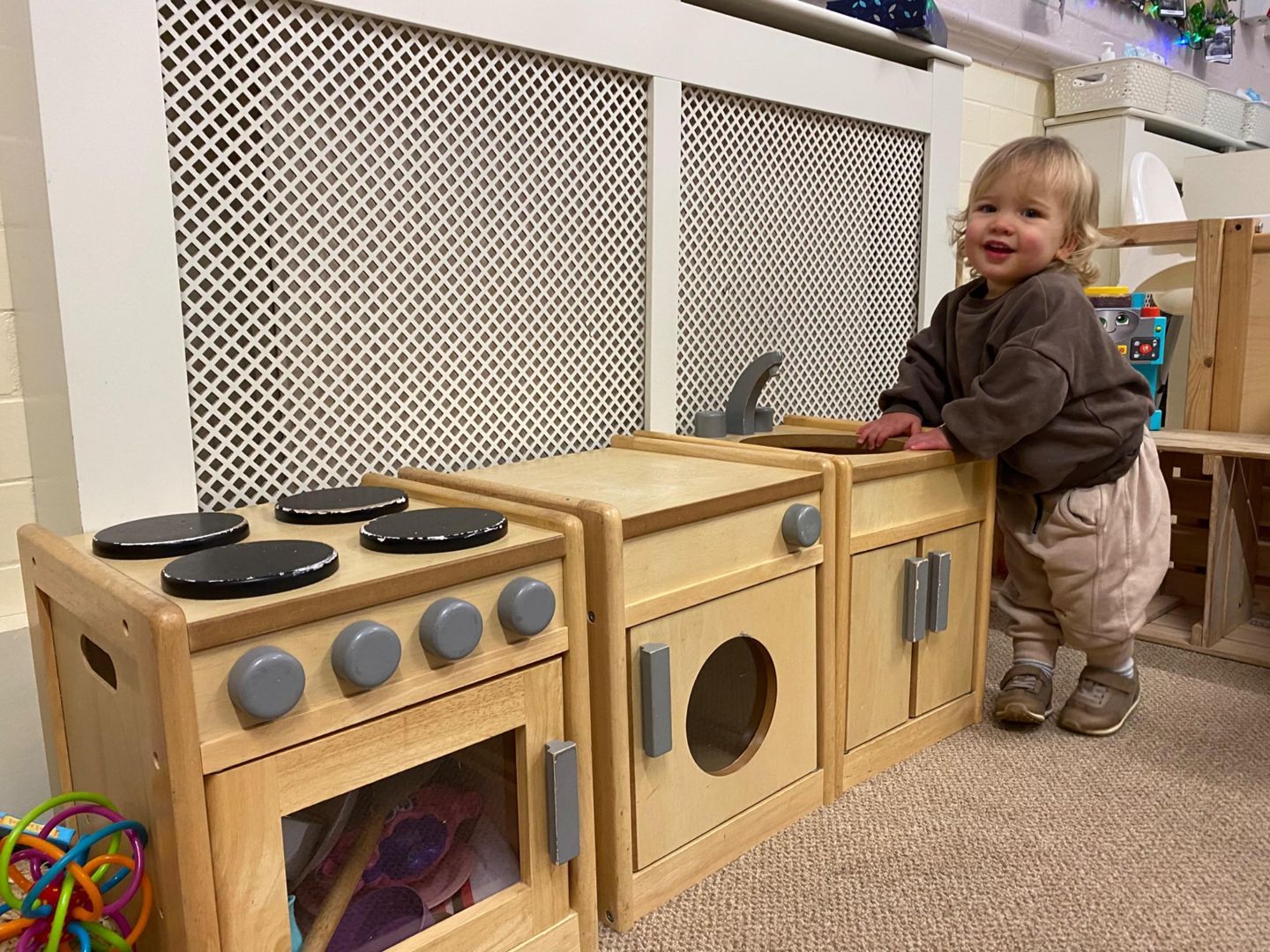 Smiling toddler at a wooden toy kitchen set, leaning on it in a room with a radiator.