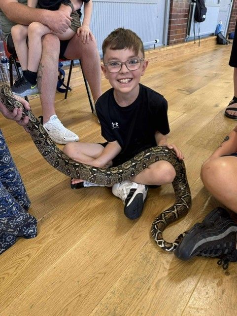 Boy smiling, holding a snake on wooden floor, wearing glasses and a black shirt.