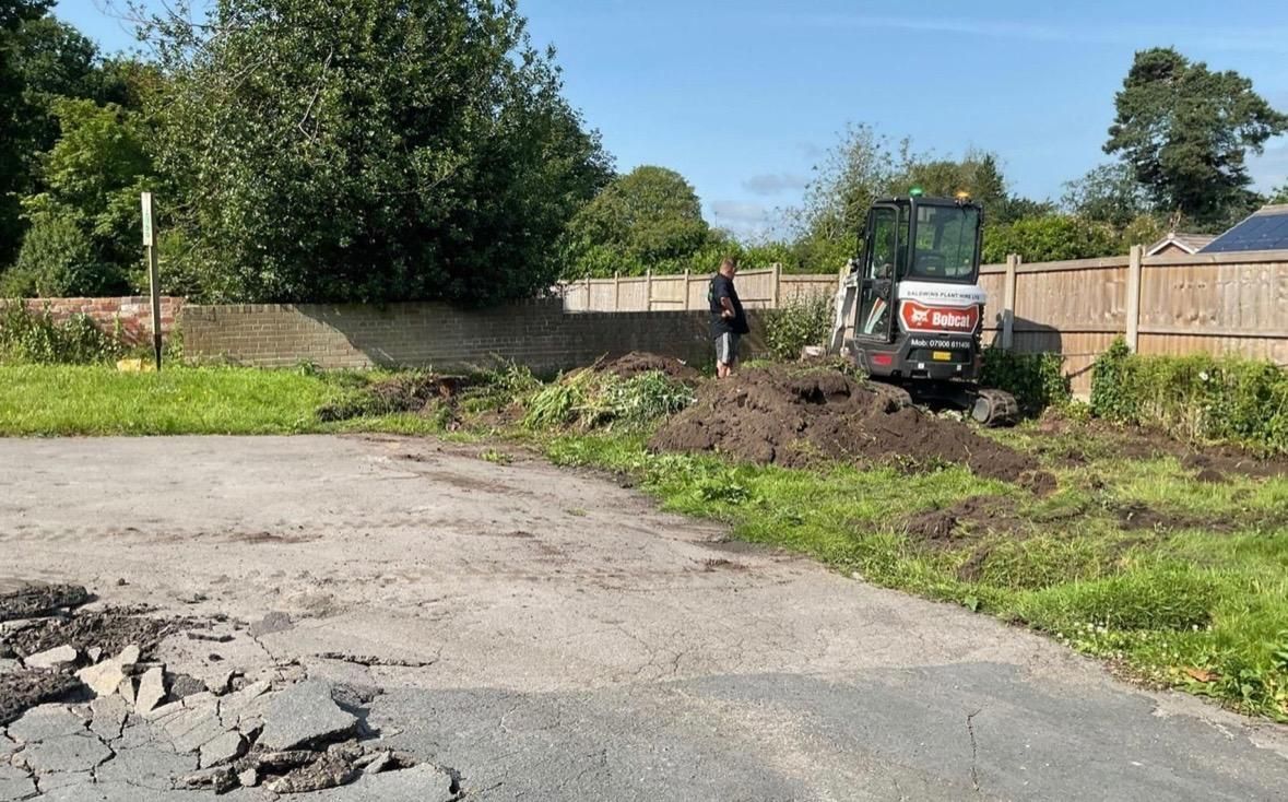A small excavator and person working in a dirt pile next to a damaged pavement.