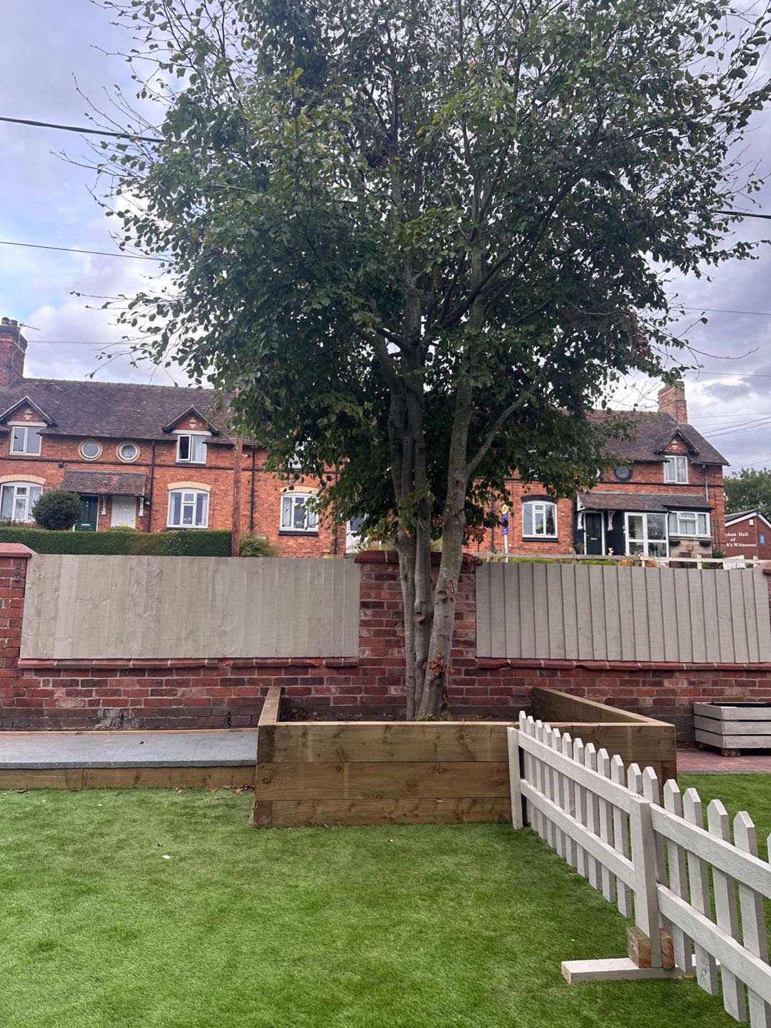 A tree in a wooden planter, surrounded by a white picket fence and a brick wall, with houses in the background.