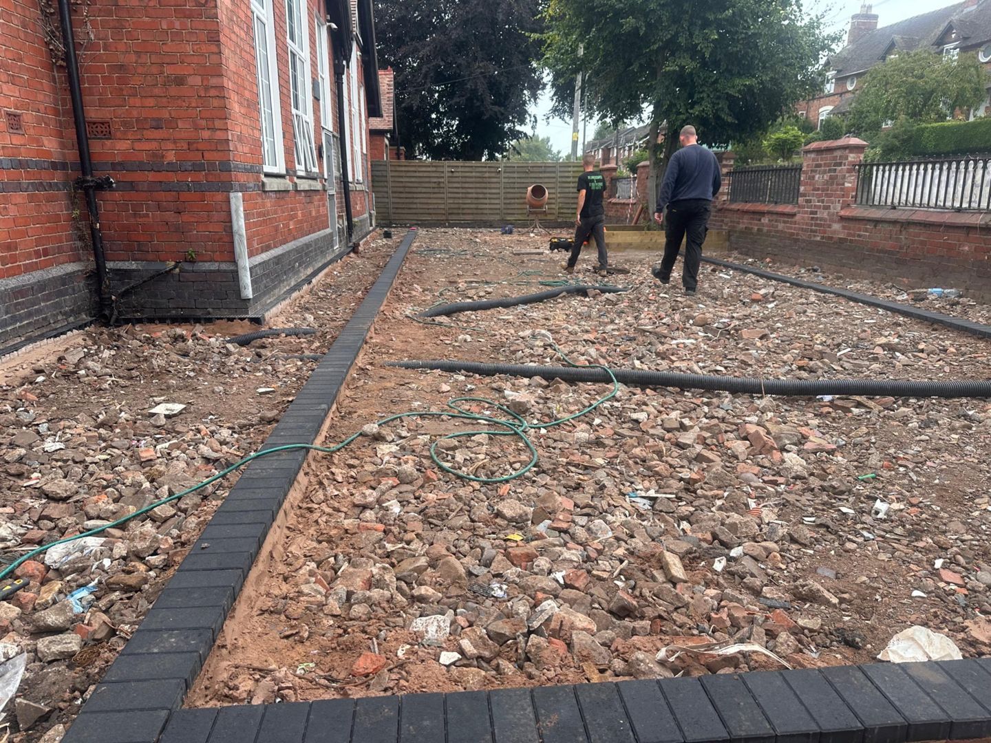 Two people working on a debris-filled yard, bordered with black paving bricks, next to a brick building.