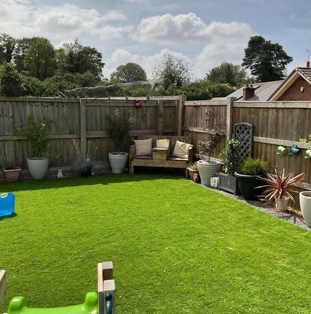 Lush green backyard with a wooden fence, bench, and potted plants. Sunny day with blue sky and clouds.