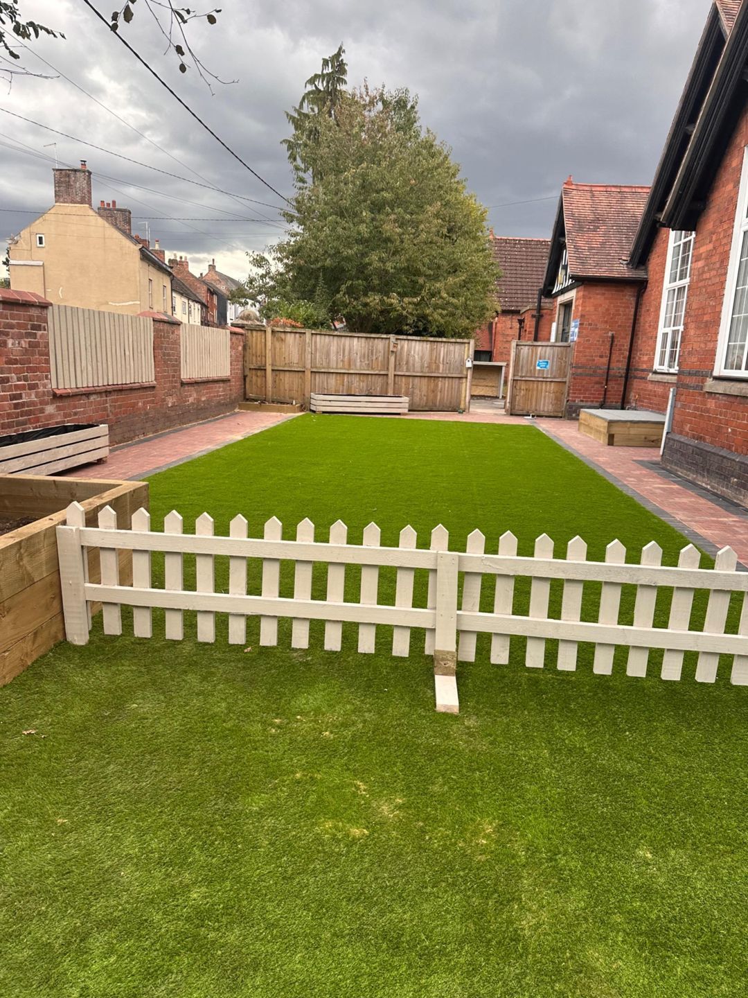 A small yard with green turf, white picket fence, and a wooden fence, with a building in the background.