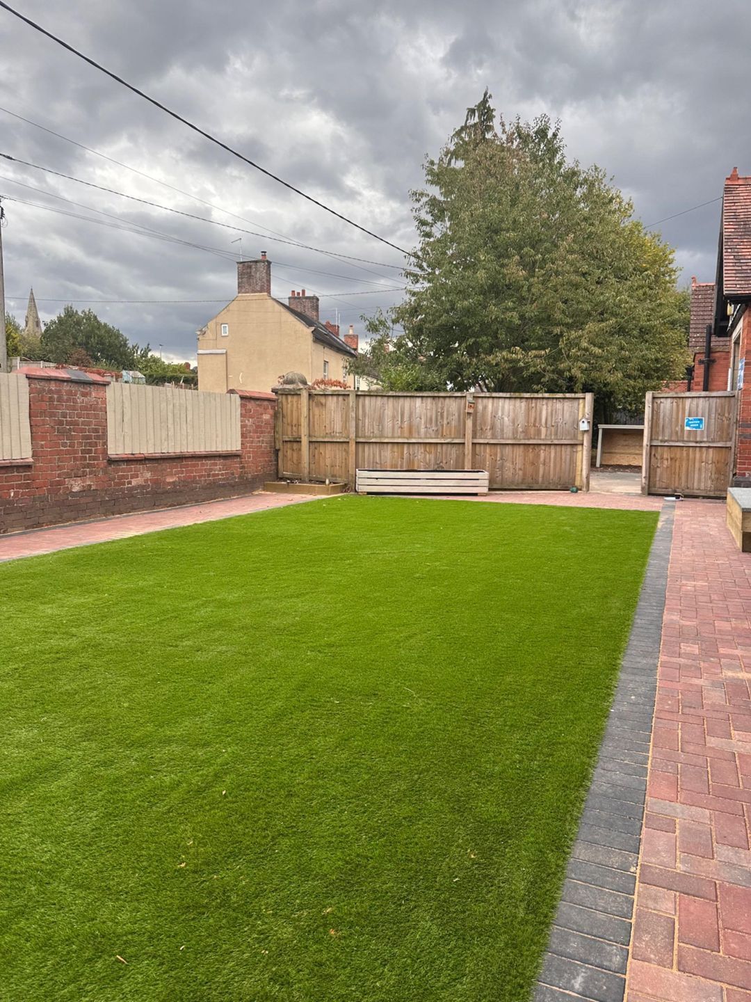 Green lawn in a backyard, bordered by brick and wooden fences, overcast sky.