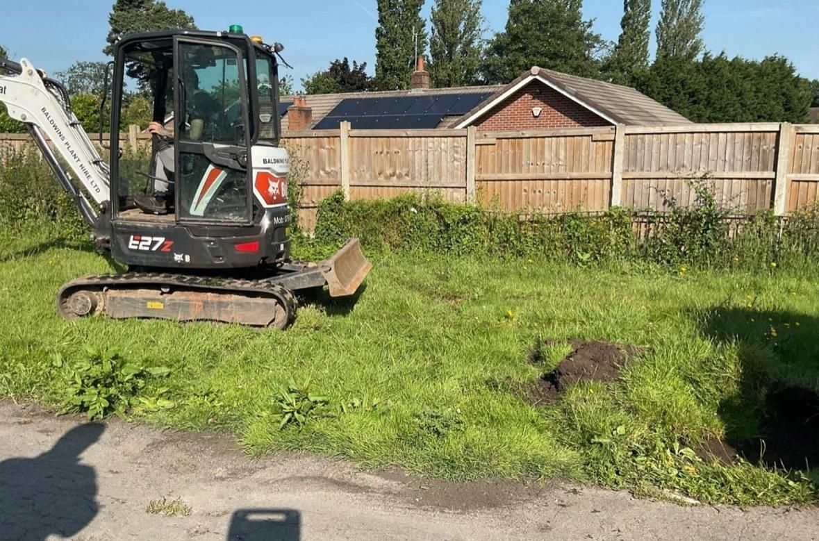 Mini excavator on grassy verge near a fence, digging earth on a sunny day.