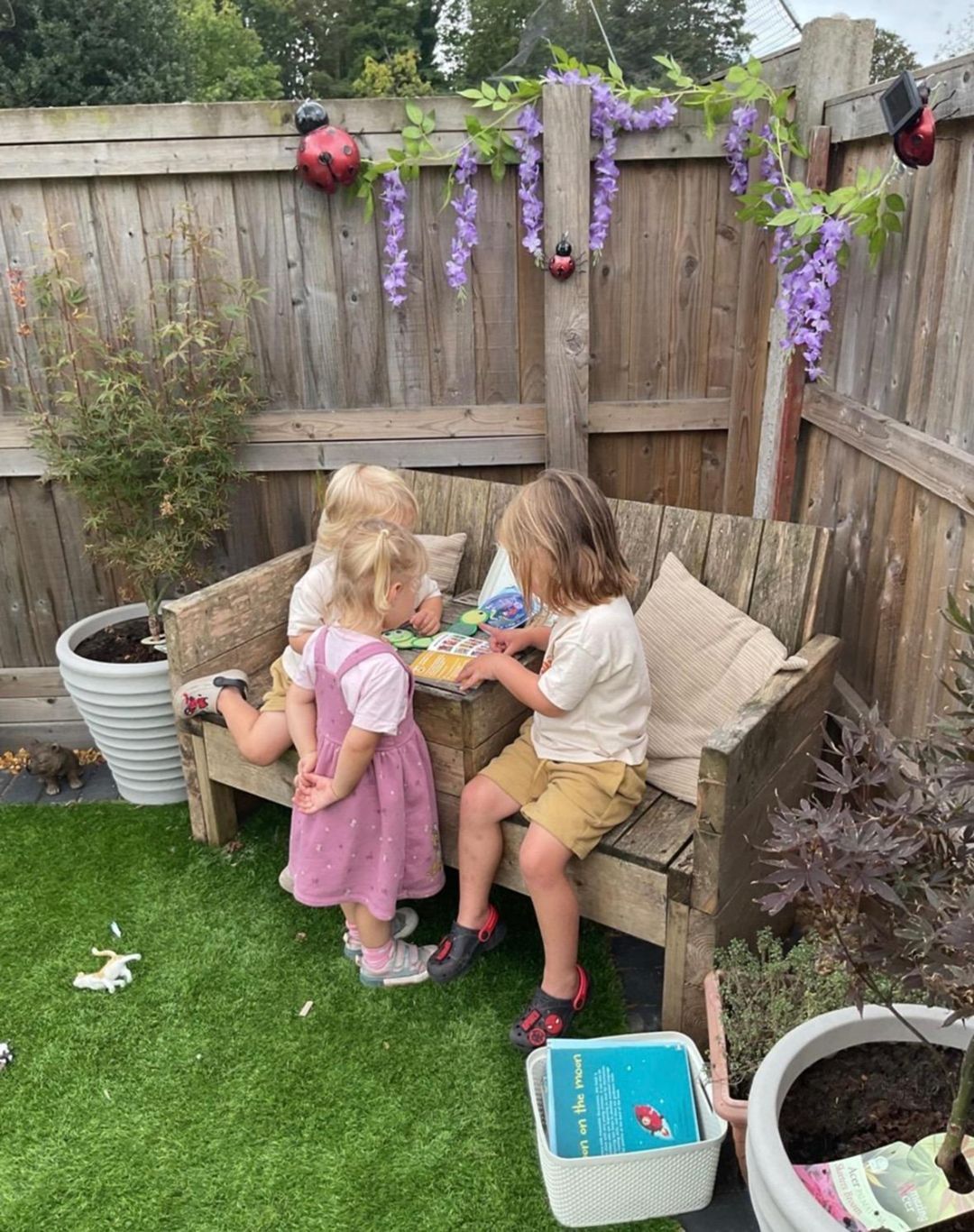 Three children gathered around a wooden bench, looking at a cake on a small table in a garden setting.