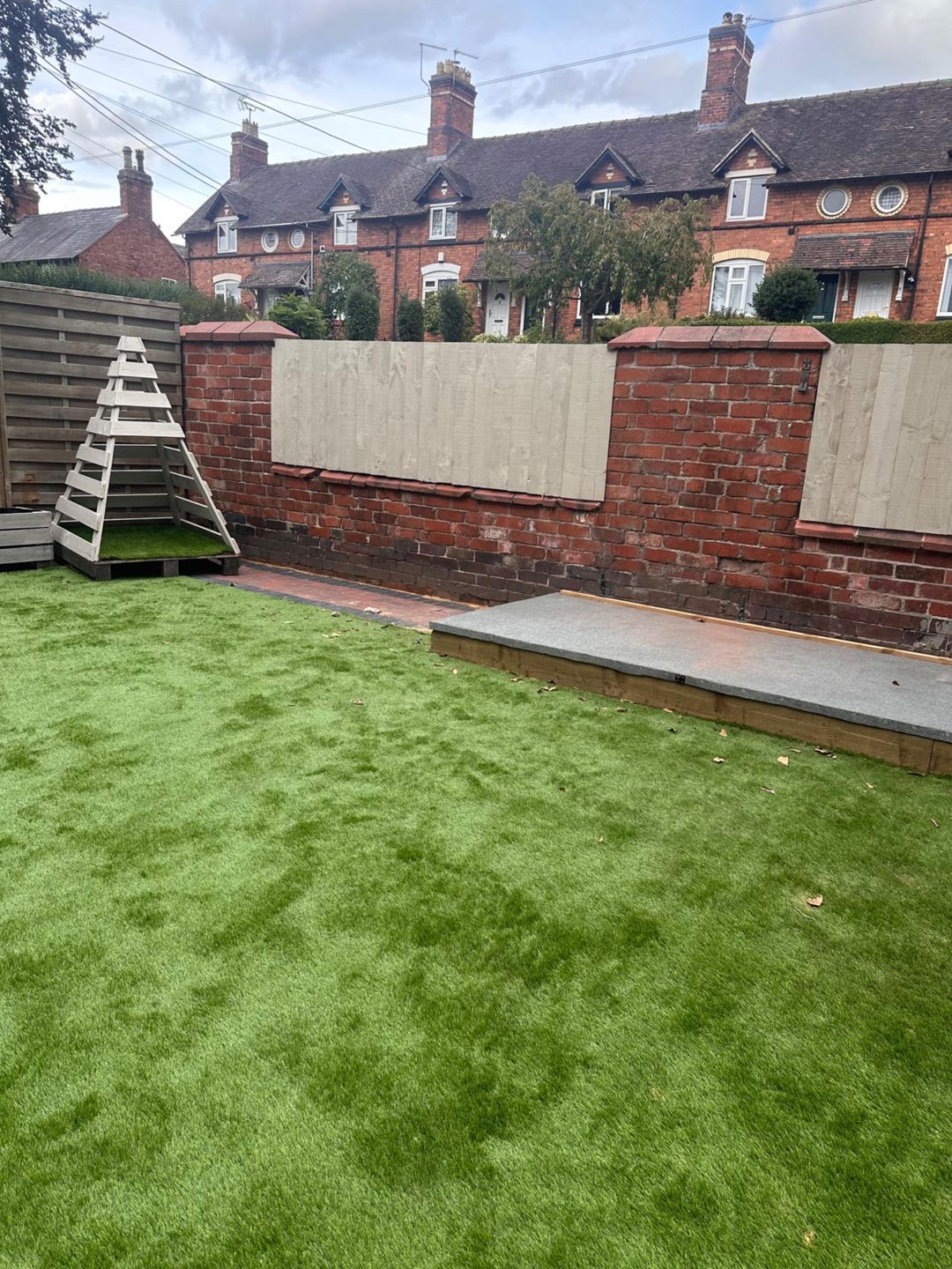 A backyard with green turf, a wooden play structure, and a red brick wall with wooden panels.