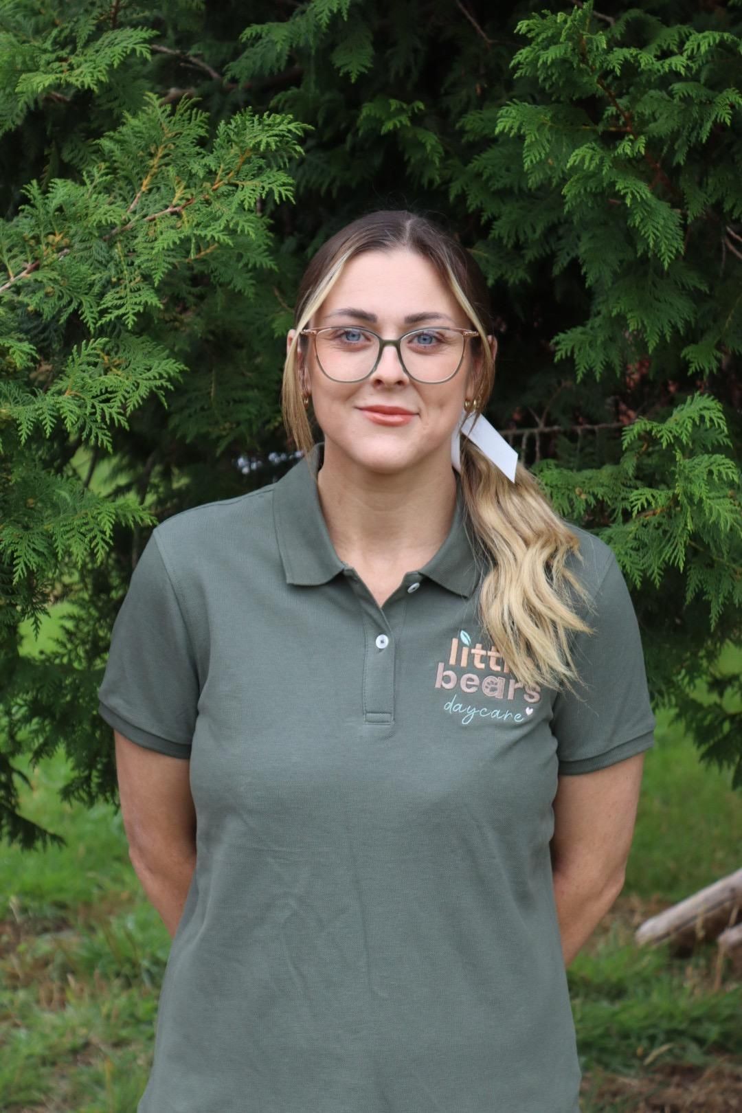 Woman in green polo shirt, glasses, and ponytail smiling outdoors, logo visible on shirt.