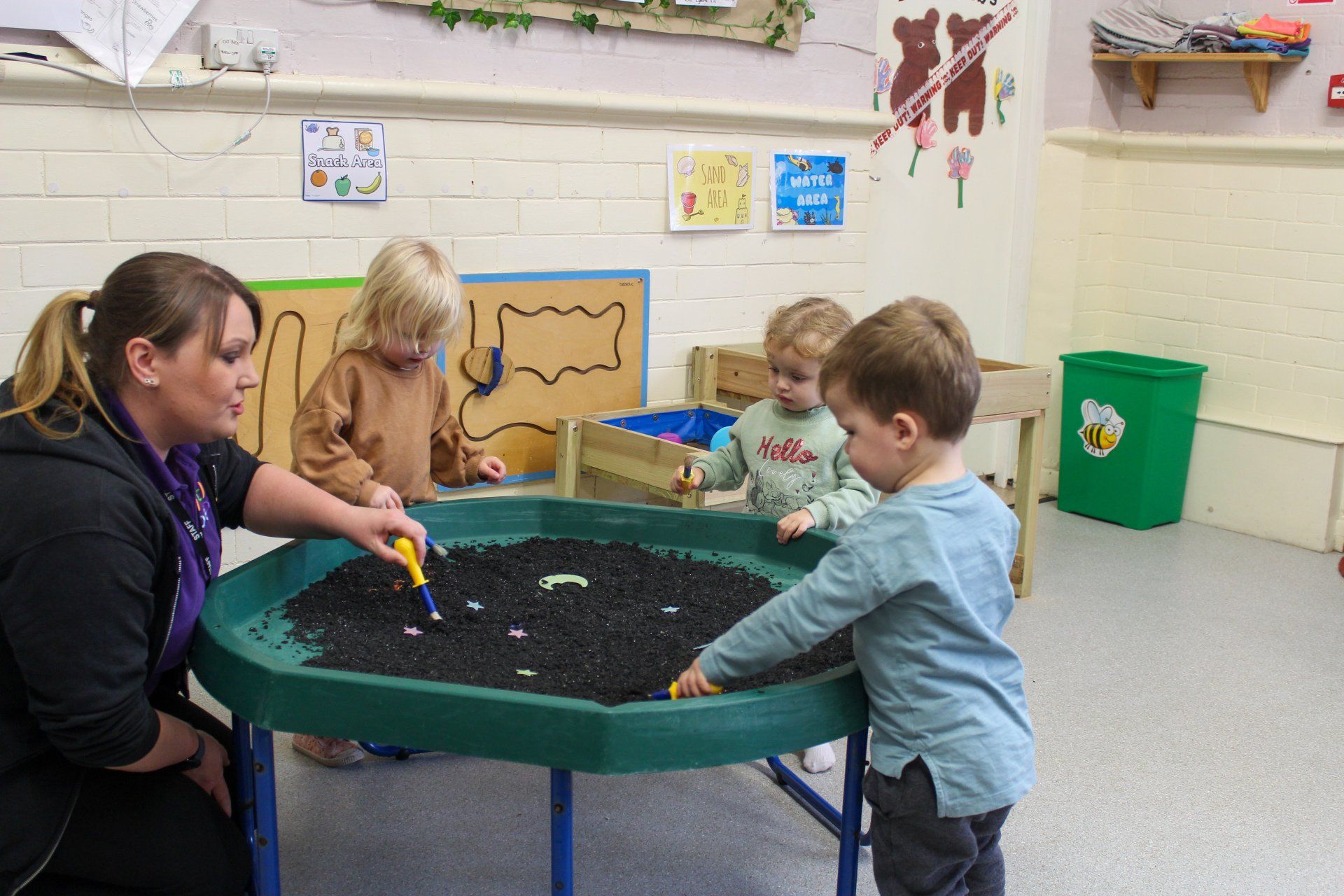 A teacher and three toddlers playing with dark beads in a green tray. Indoors, learning activity.