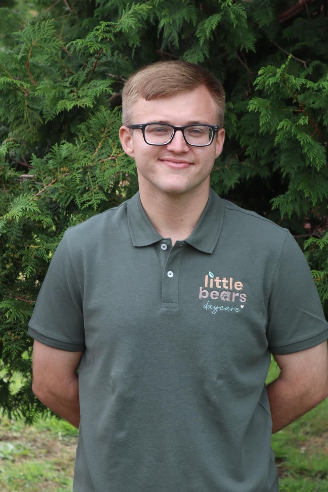 Man wearing glasses and green polo shirt with logo, arms behind his back, standing in front of greenery.