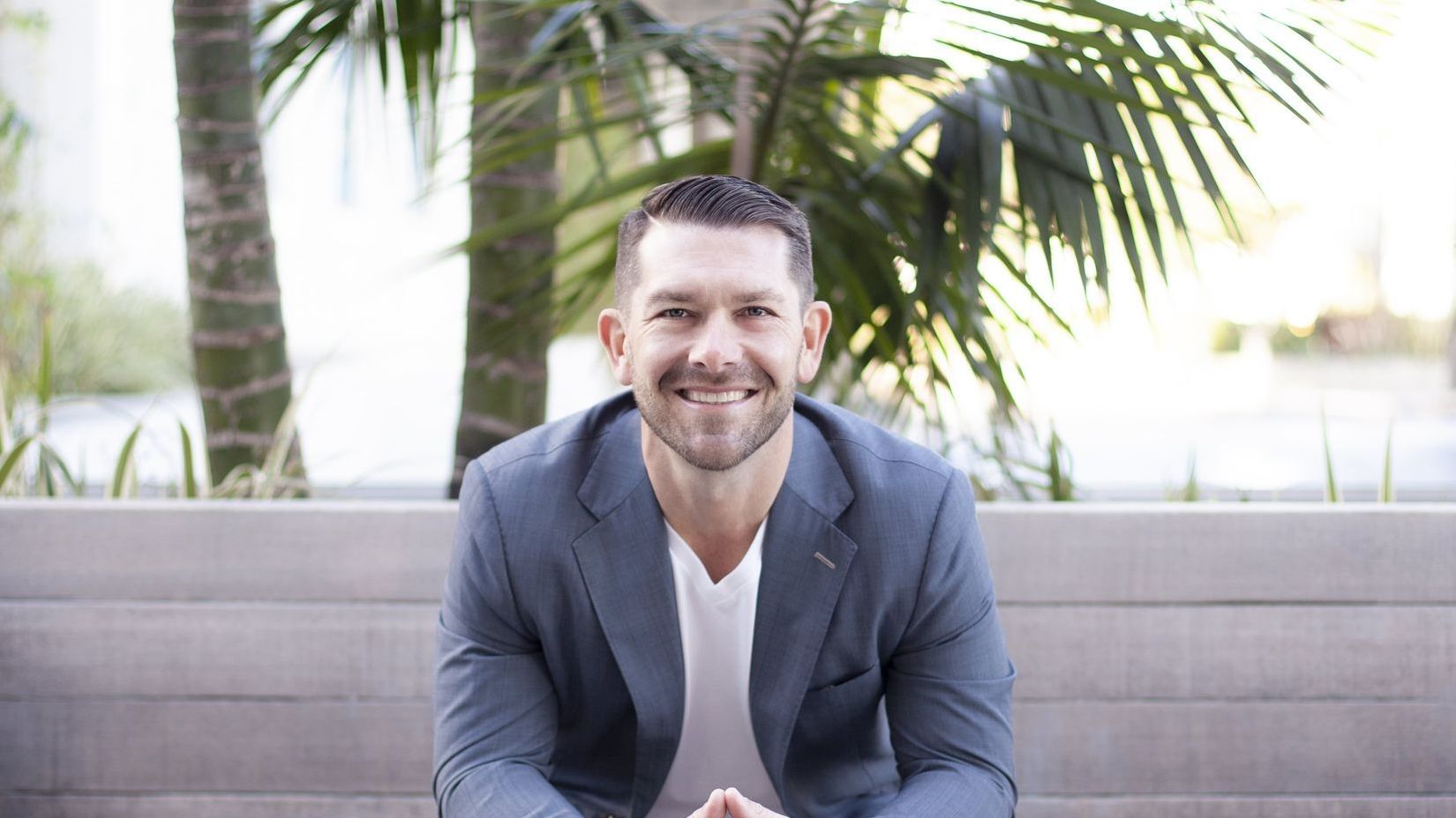 Smiling man in a gray blazer sitting outdoors on a bench with palm trees behind him