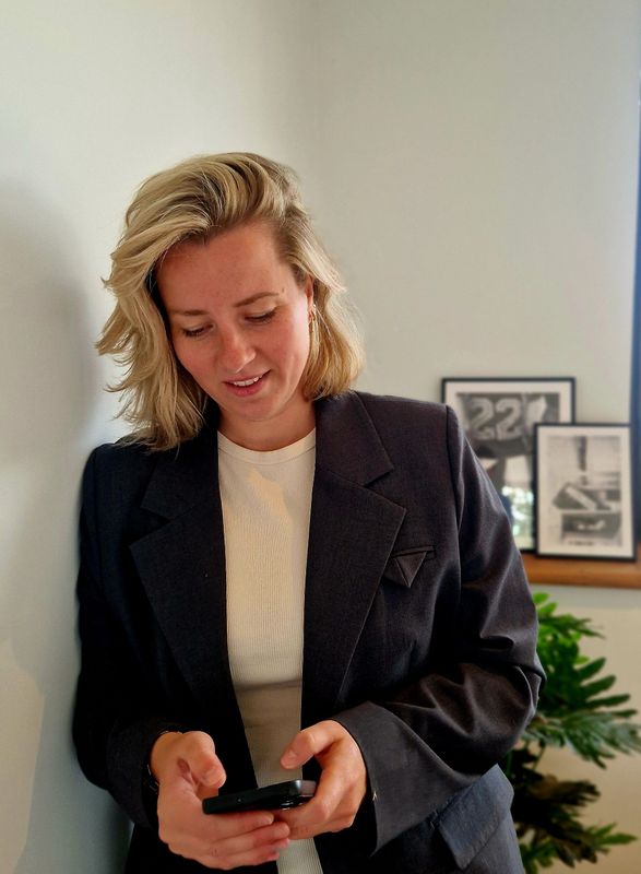 Vrouw in een donkere blazer kijkt binnen naar haar telefoon, naast ingelijste kunst en een plant.
