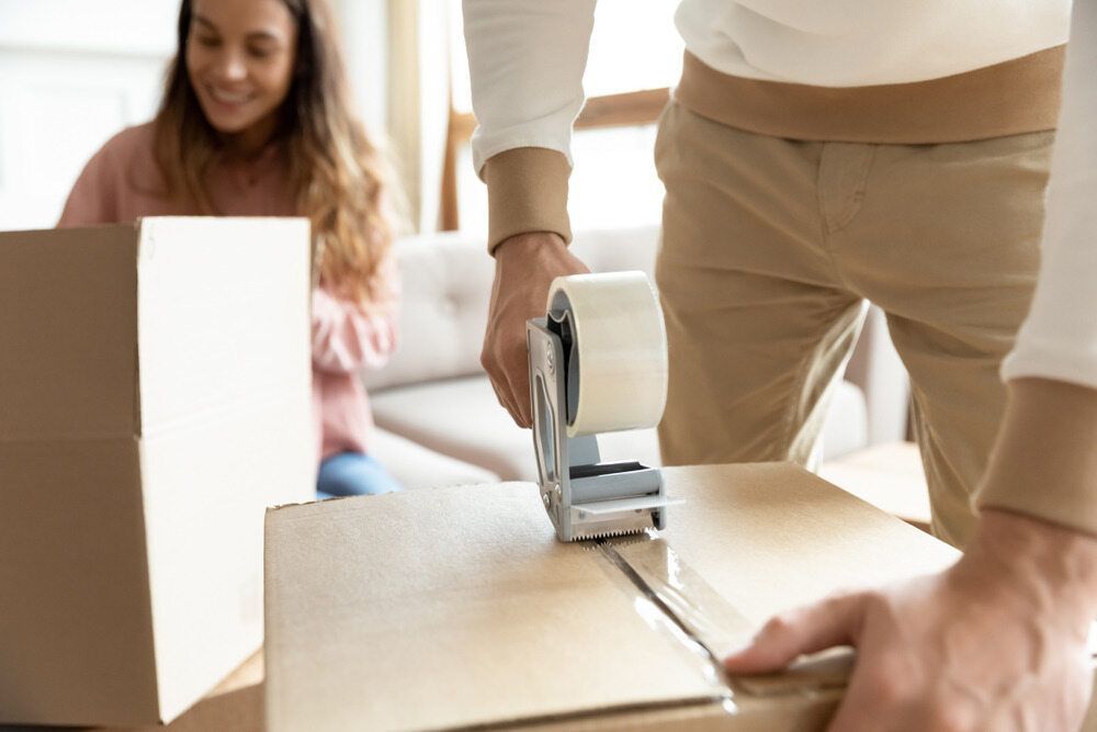A Man is Taping a Cardboard Box With Tape — Johns Removals Newcastle In Cessnock, NSW