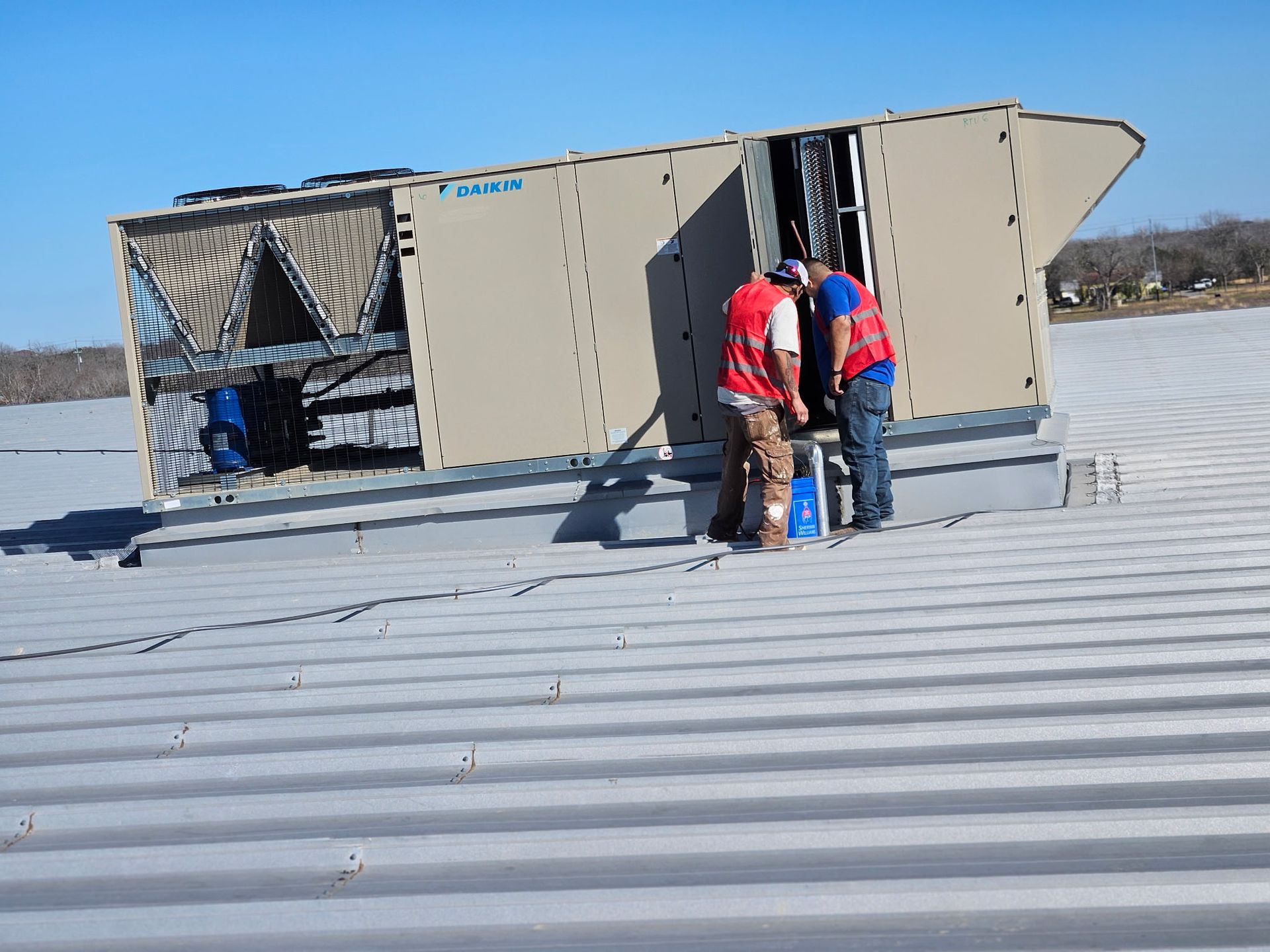 Two workers in red vests near an HVAC unit on a rooftop.
