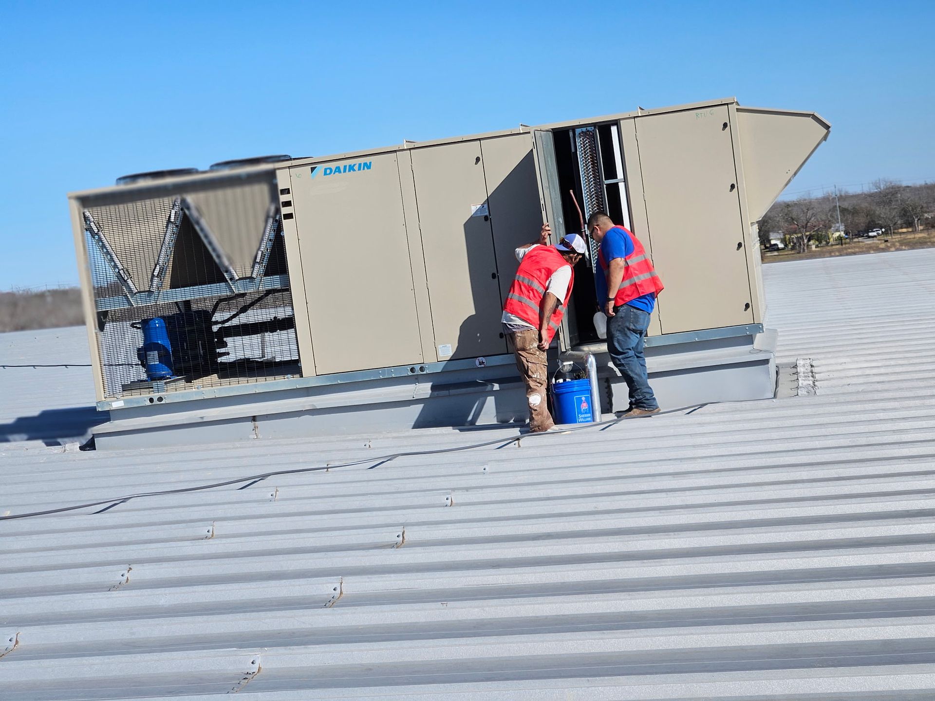 Two workers in safety vests near an HVAC unit on a rooftop.