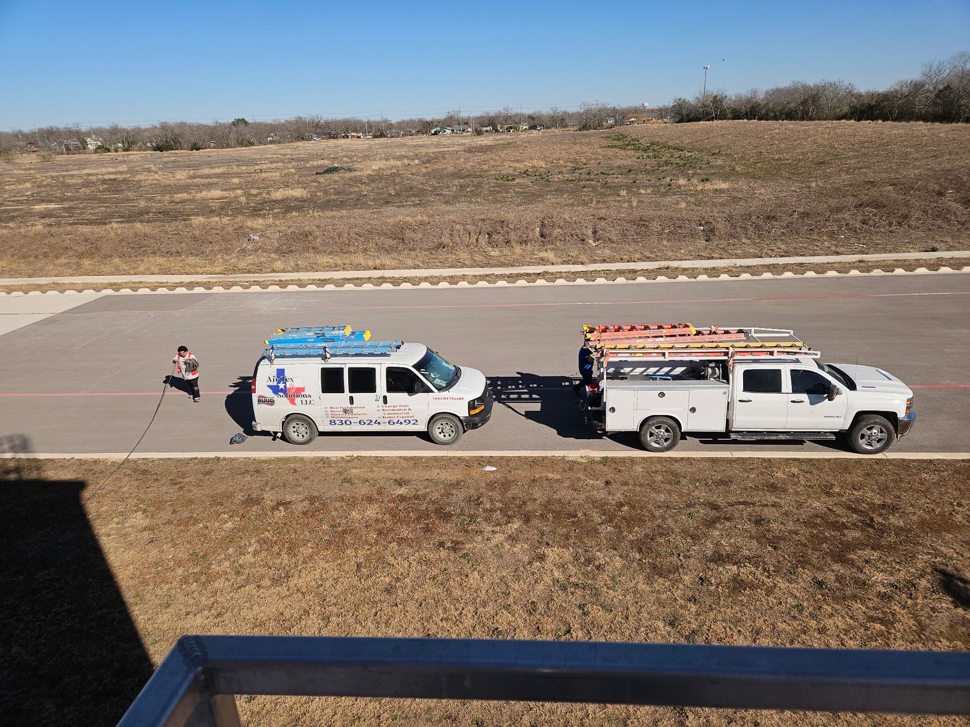 Two work trucks with ladders parked on asphalt. A person stands near them on a sunny day.