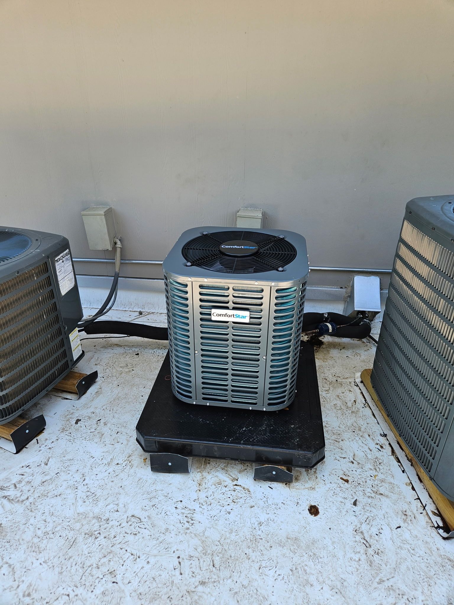 Central air conditioning unit on a rooftop platform, flanked by two other units, against a light gray wall.