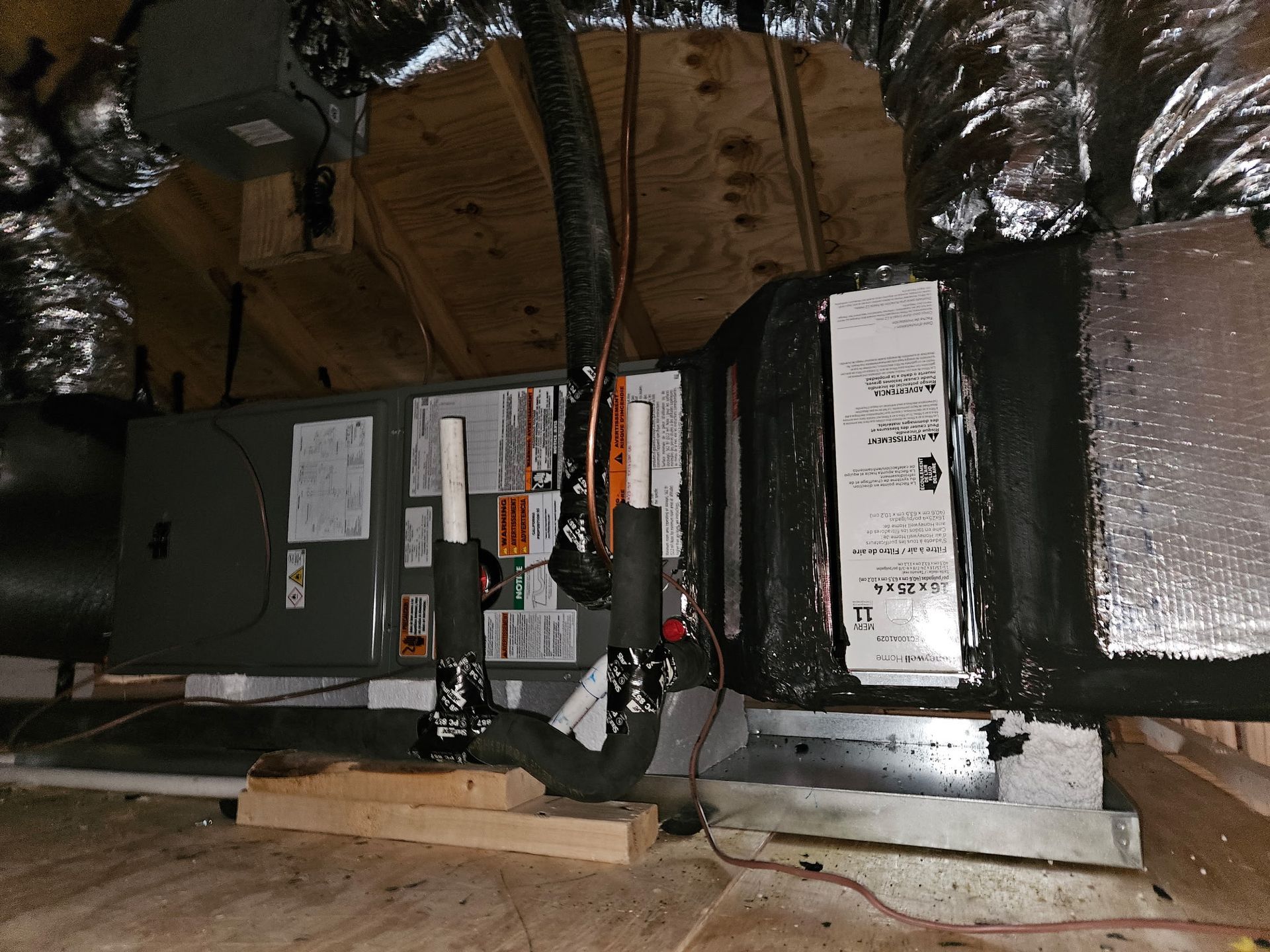 HVAC system in an attic. Gray metal unit with black ductwork, silver insulation, and wood supports.