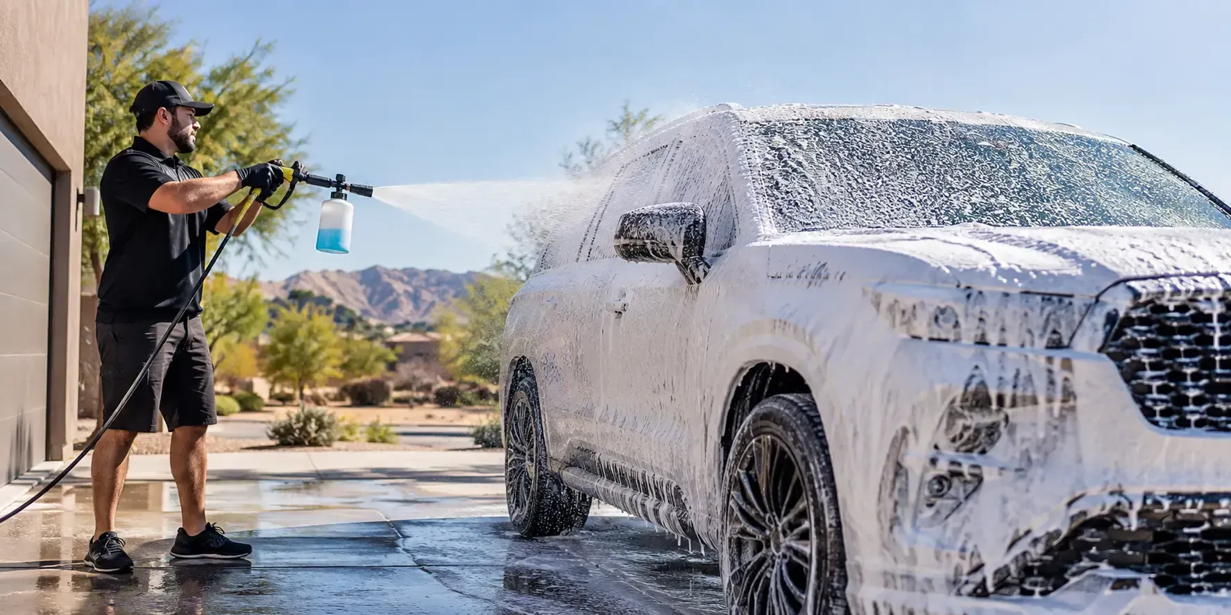 Professional car detailer using a foam cannon to cover a white SUV in thick snow foam, preparing to safely remove desert dust.