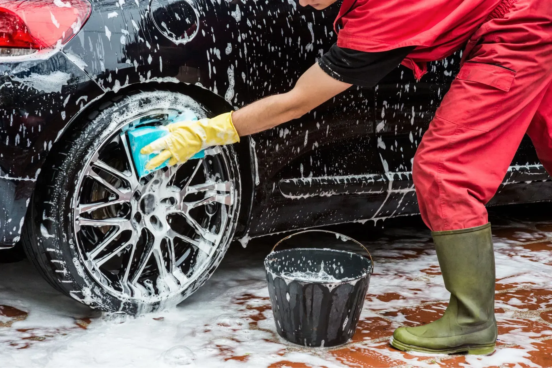 Mobile Detailing - Person washing a black car, using a sponge and soapy water, next to a bucket.