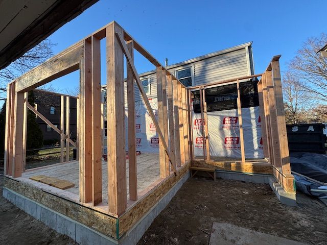 A wooden house addition in the framing stage, featuring unfinished wall studs on a foundation against an existing home.