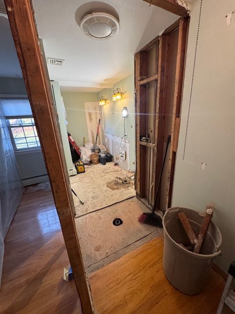 A doorway view of a room under renovation with exposed wooden wall studs, tools, and a trash bin on a wooden floor.