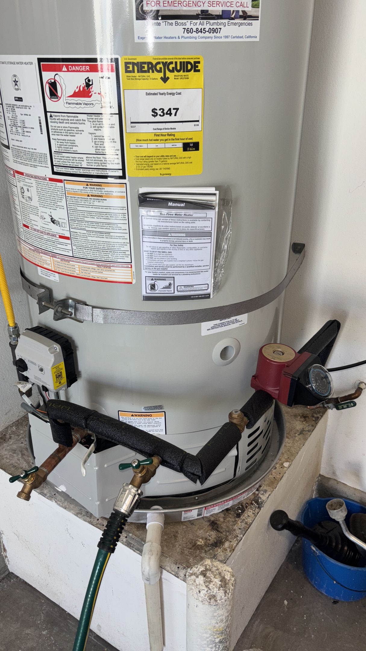 Water heater with multiple labels and connected pipes in a utility room, with a blue bucket beside it