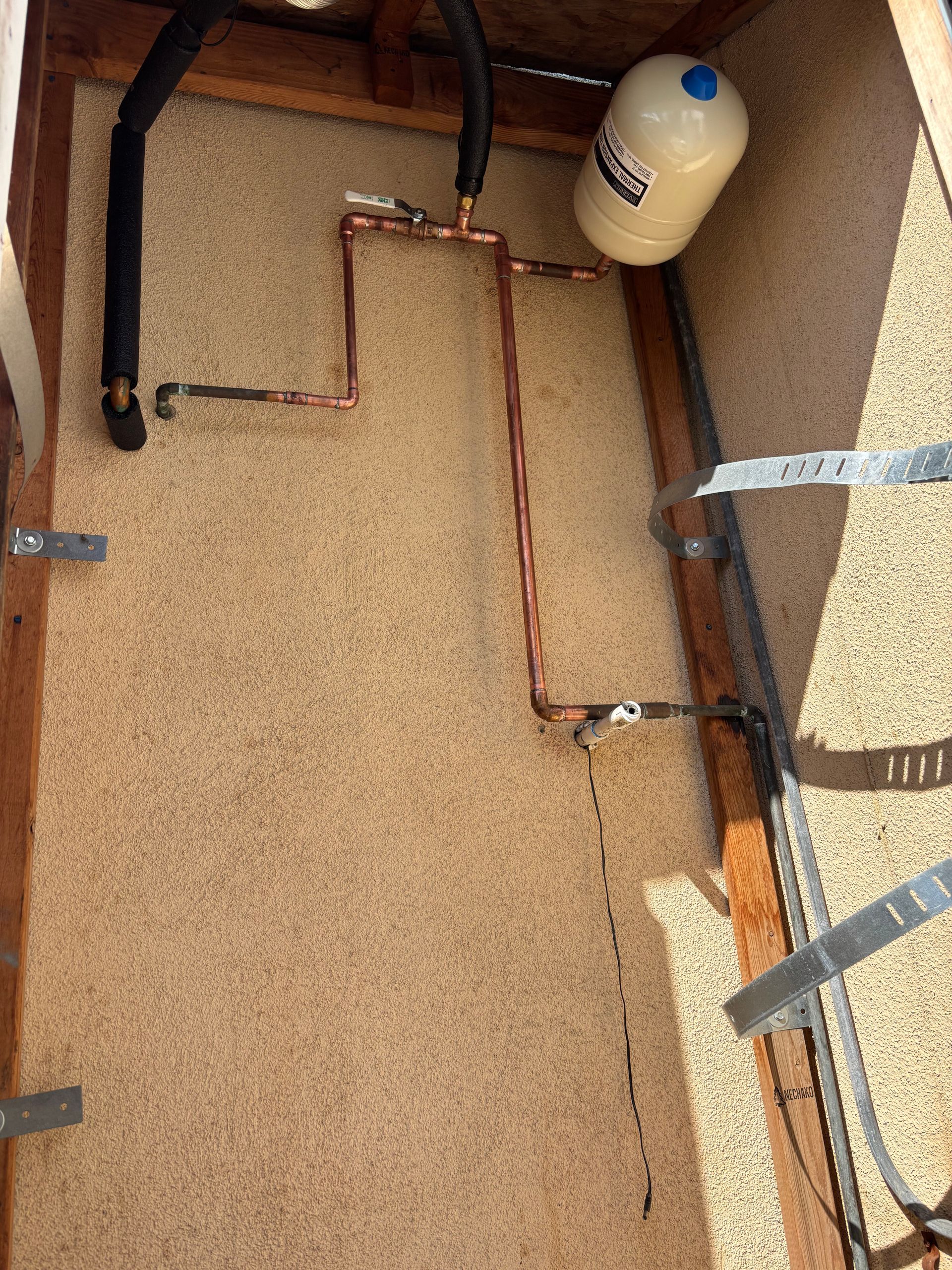 Open utility cabinet with copper pipes, black hoses, and a white tank against a tan wall.