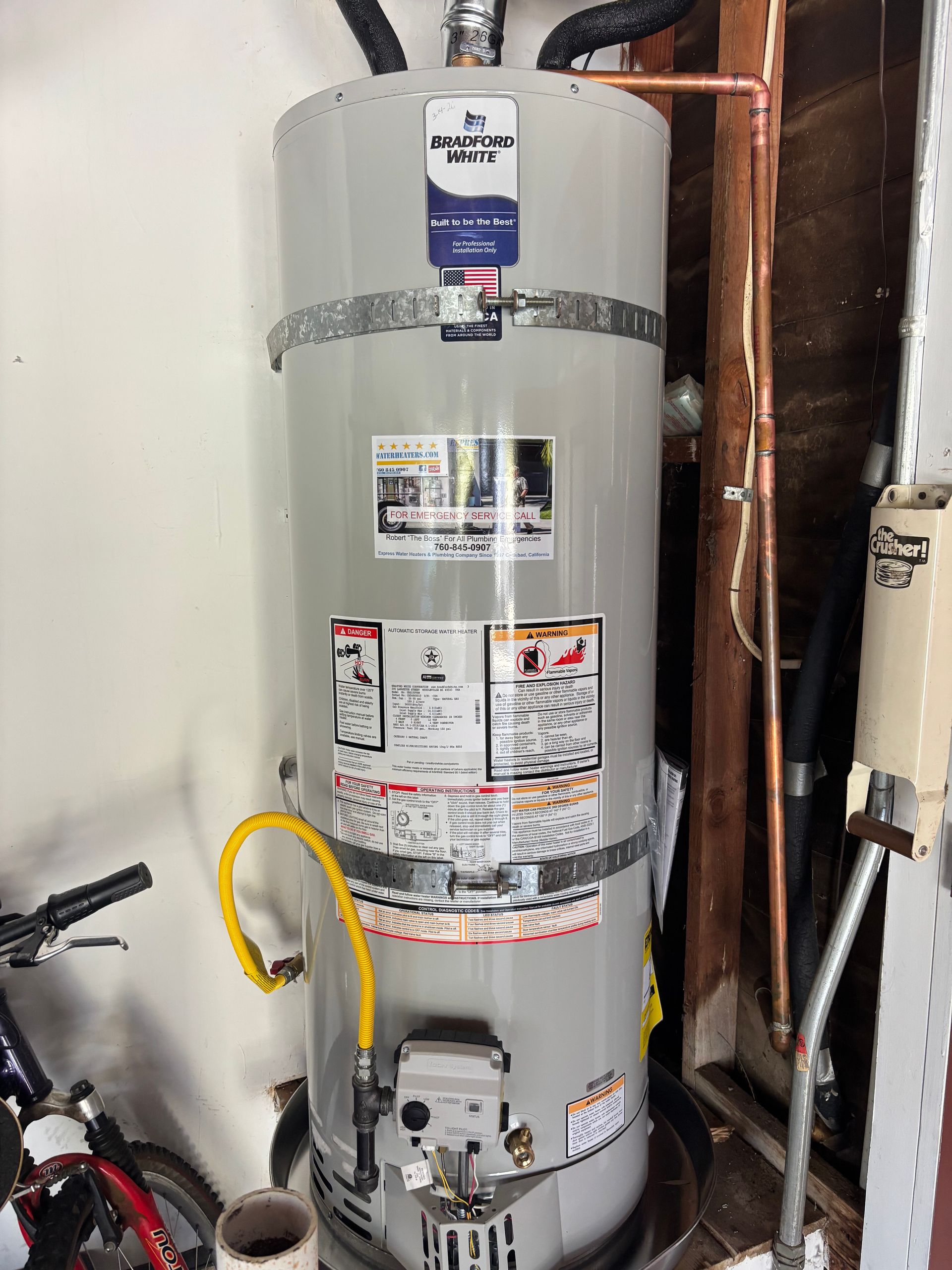 Gas water heater in a utility closet with pipes and labels, mounted beside plumbing and a wall.