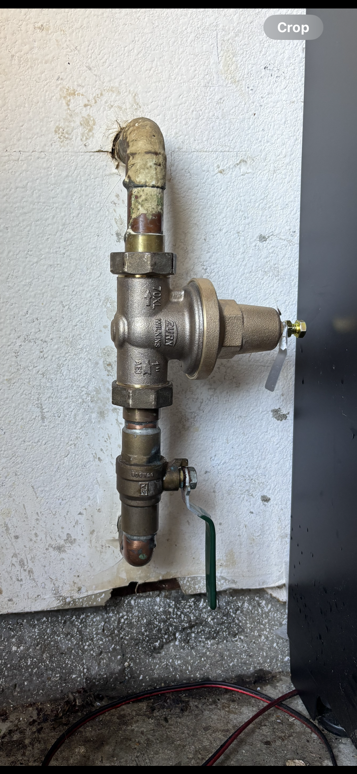 Old metal plumbing valve and pipe assembly mounted on a rough wall with a hose outlet and green tube.