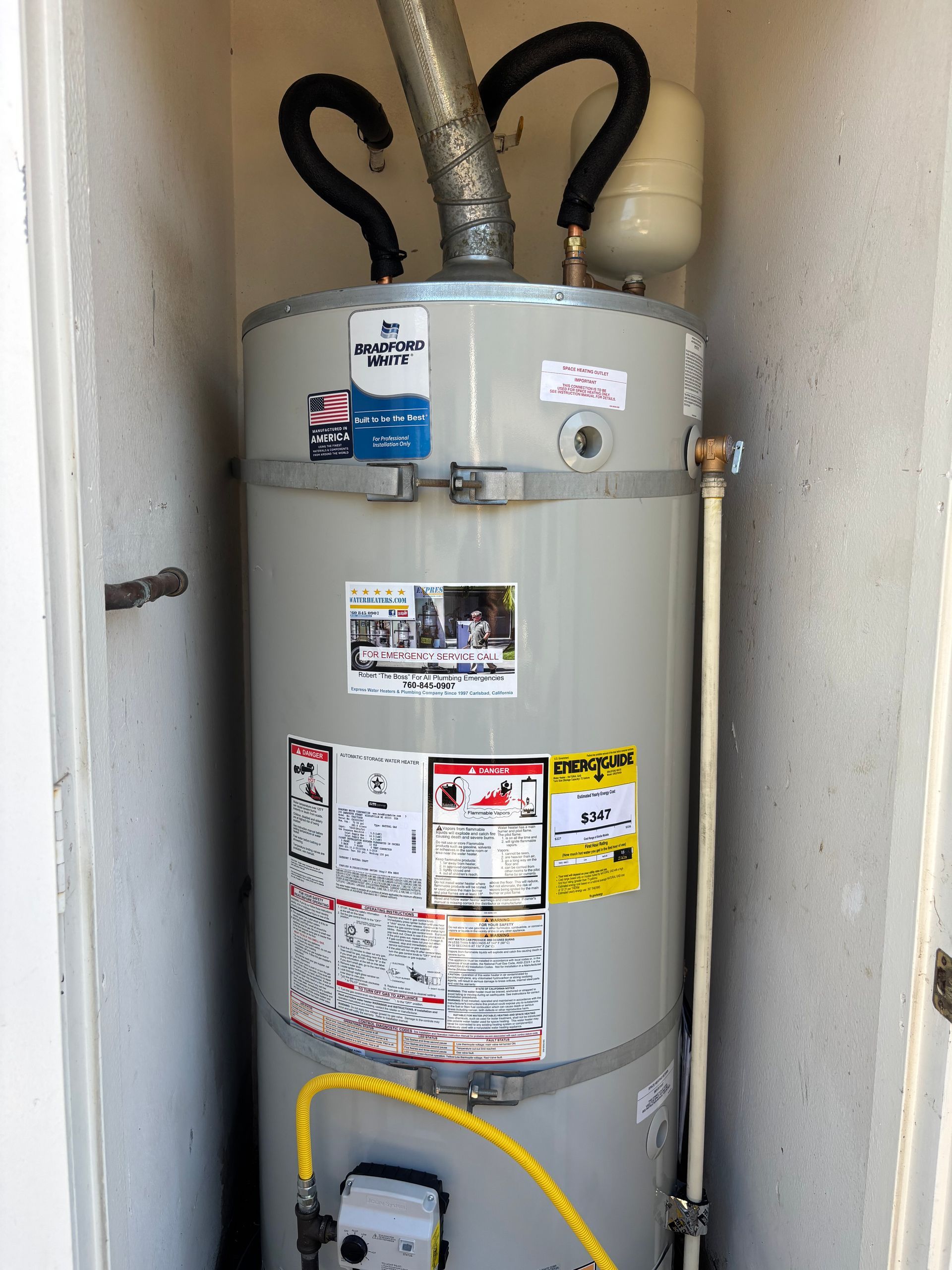 Tall gray water heater in a closet with pipes and labels, front view.