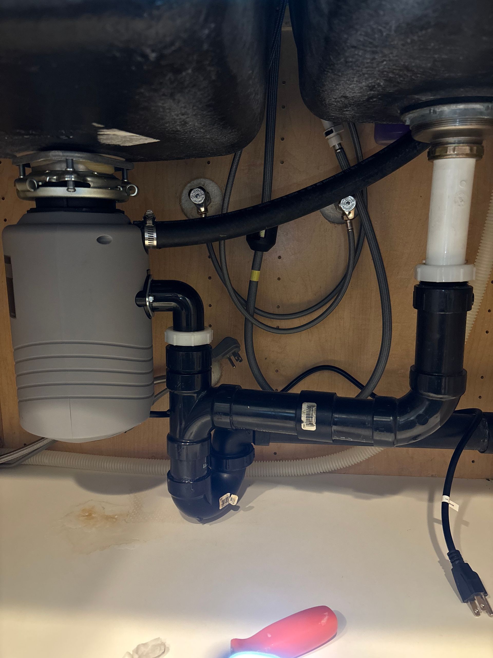Under-sink plumbing with black drain pipes, gray disposal unit, and a red-handled tool below.