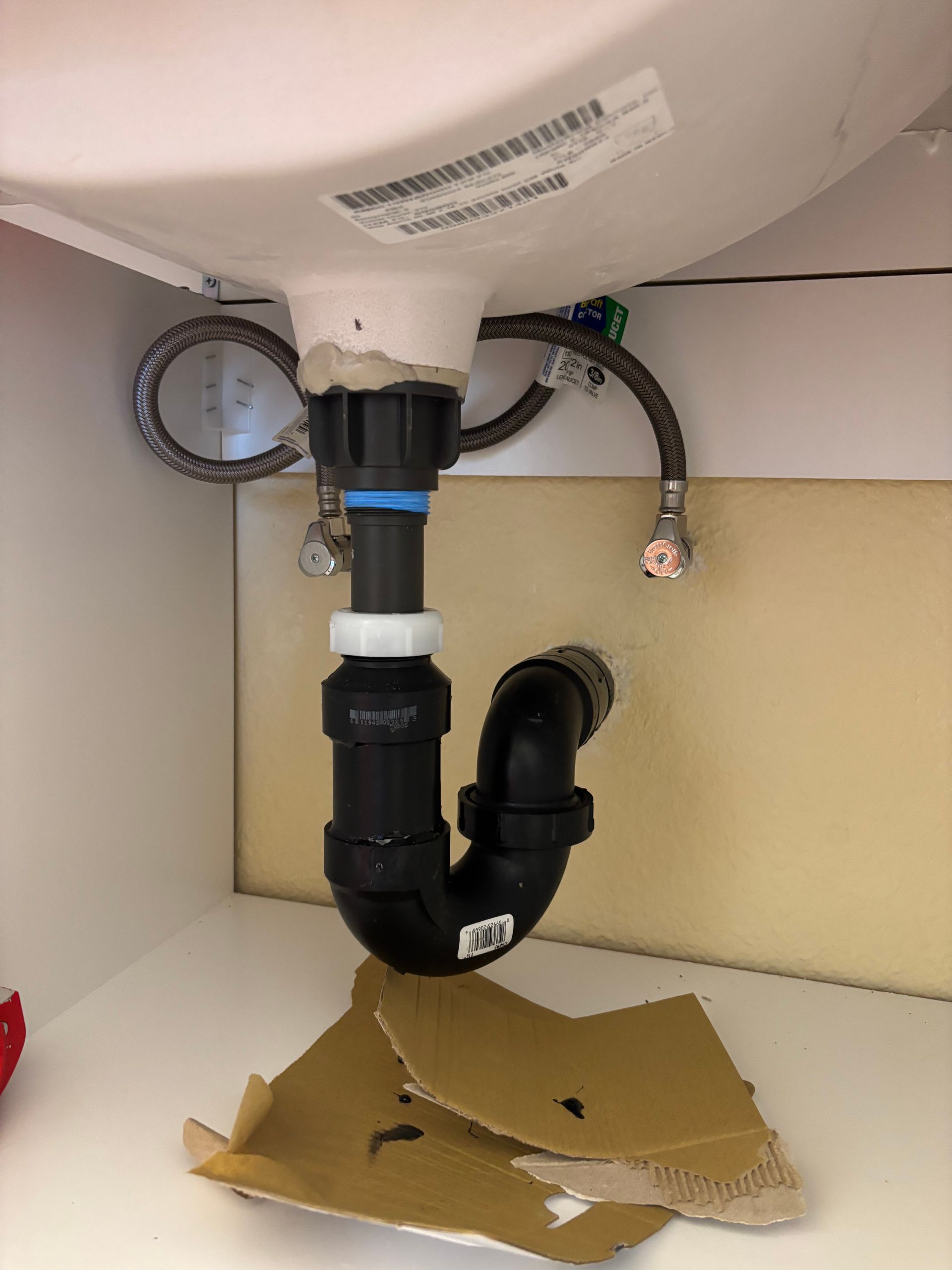 Under-sink plumbing with black drain pipe, loose cardboard on cabinet floor, and visible water supply lines