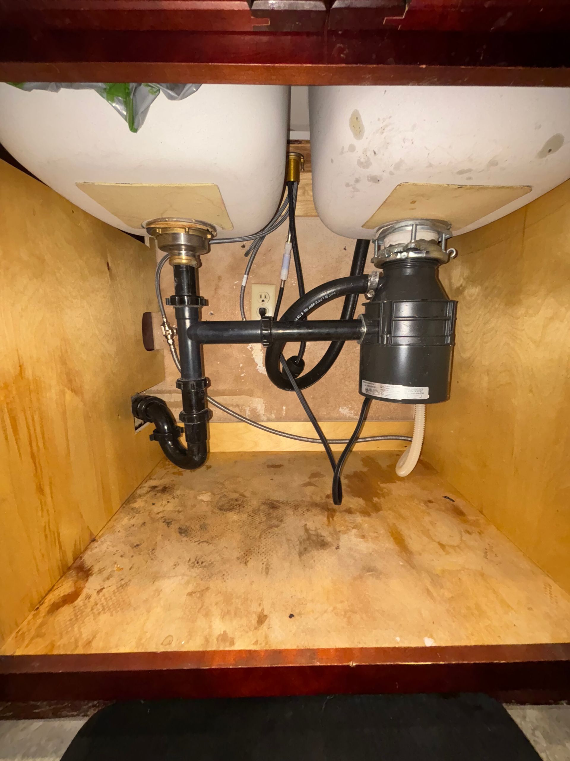Under-sink cabinet with exposed plumbing, two basins, and a dirty stained floor