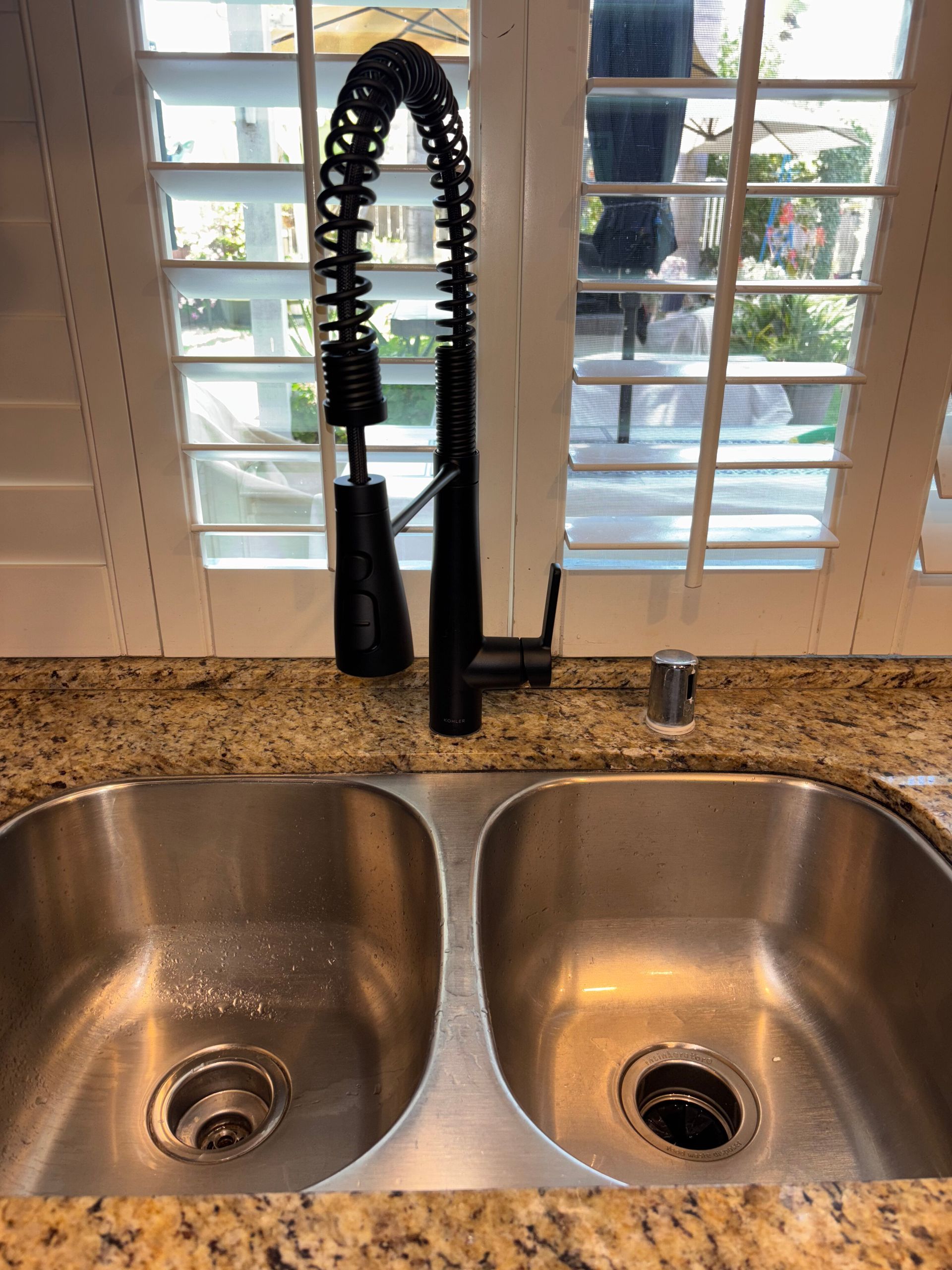 Double kitchen sink under a window with shutters and a black spring faucet.