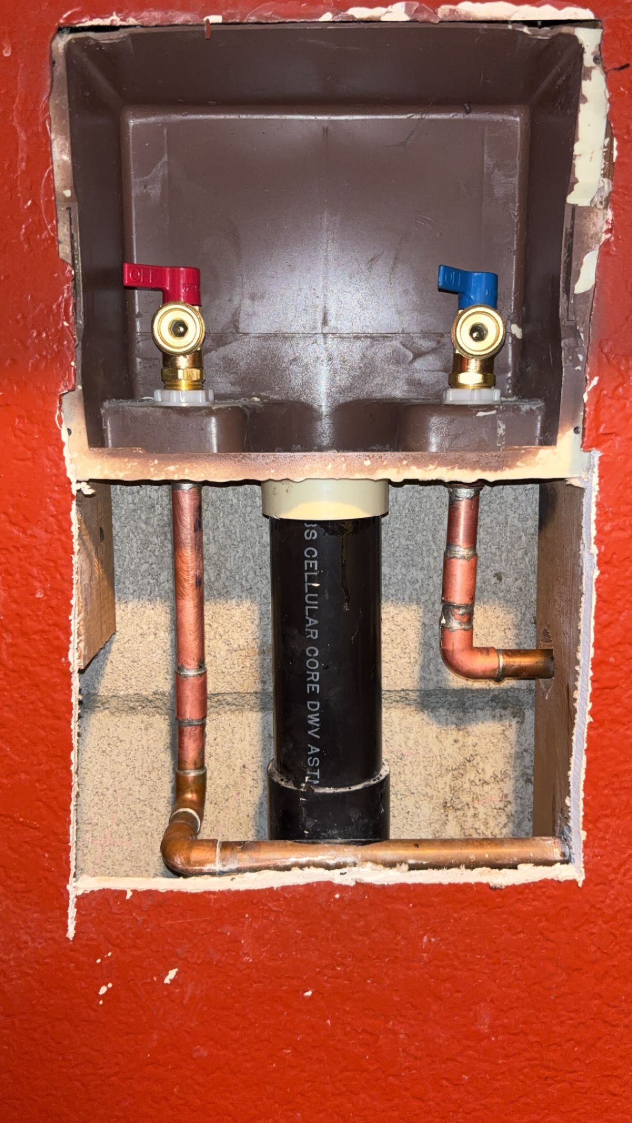 Wall-mounted plumbing access panel with red and blue valve handles and exposed copper pipes.