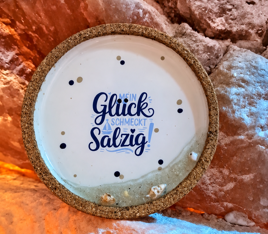 Rundes weiß-goldenes Schild vor rosa Felsen mit der Aufschrift „Glück schmeckt salzig“