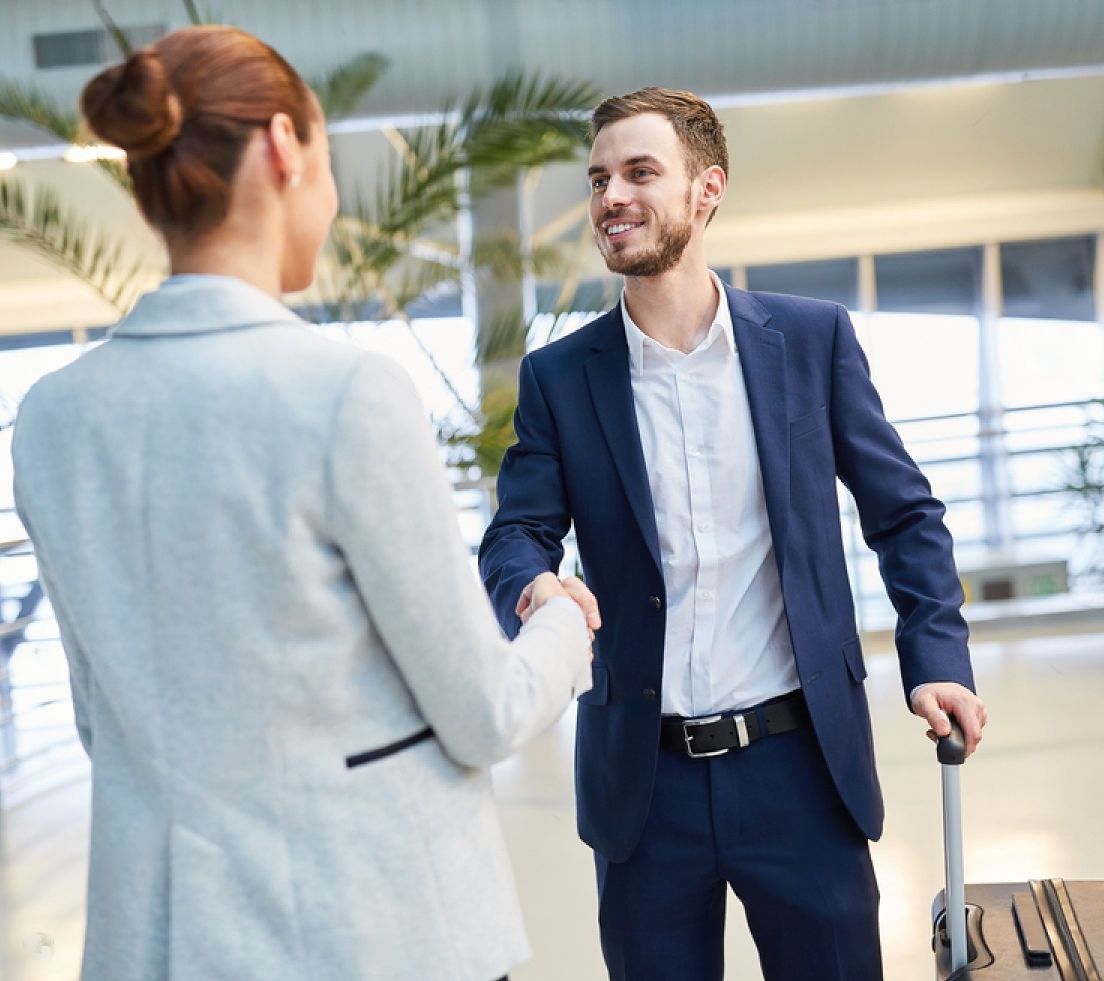 Woman and man shaking hands in an airport, man smiling, rolling luggage.