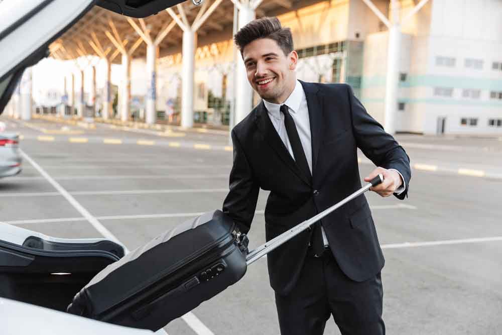 Man in suit smiling, loading luggage into a car trunk outside an airport.