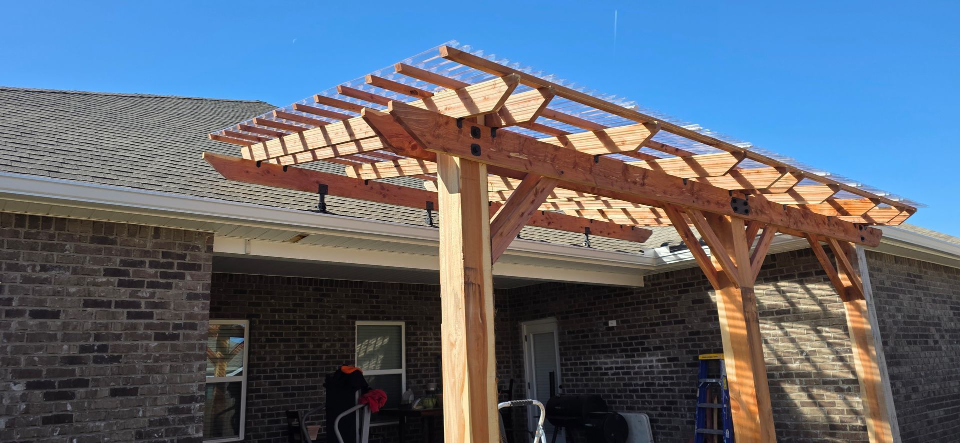 A wooden pergola with a clear corrugated roof attached to the side of a brick house under a bright blue sky.
