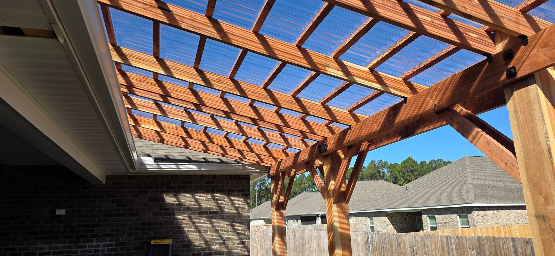 A wooden pergola attached to a brick house on a sunny day, featuring an open-slat roof design.