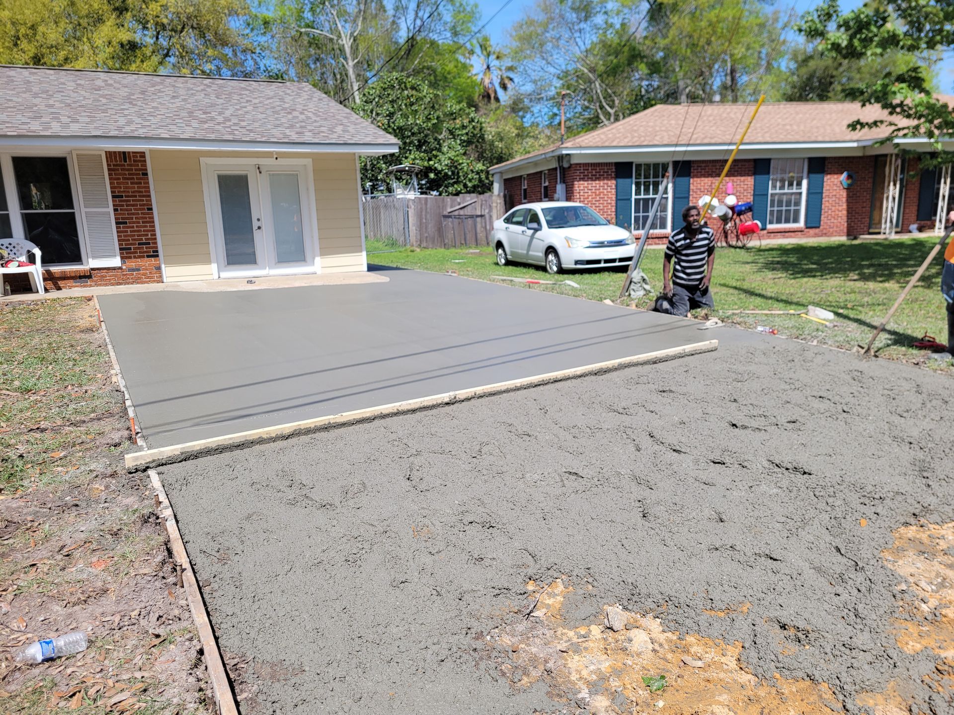 A worker finishes a new concrete patio slab extending from a residential house into a yard.